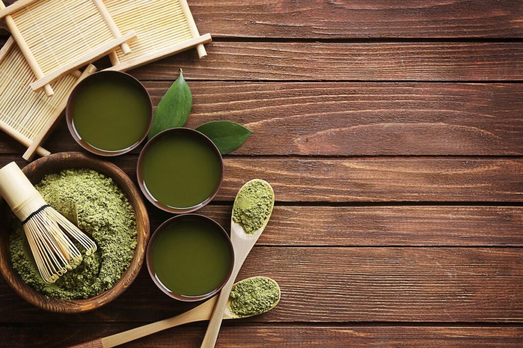 Matcha-Pulver in Schalen, aufgeschlagener Matcha-Tee und Bambusbesen auf einem Holztisch