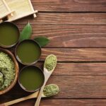 Matcha-Pulver in Schalen, aufgeschlagener Matcha-Tee und Bambusbesen auf einem Holztisch