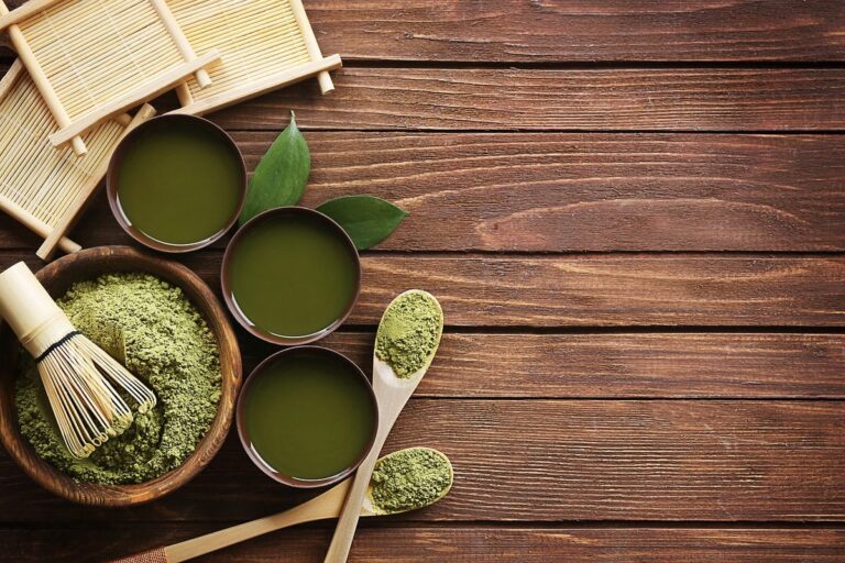 Matcha-Pulver in Schalen, aufgeschlagener Matcha-Tee und Bambusbesen auf einem Holztisch
