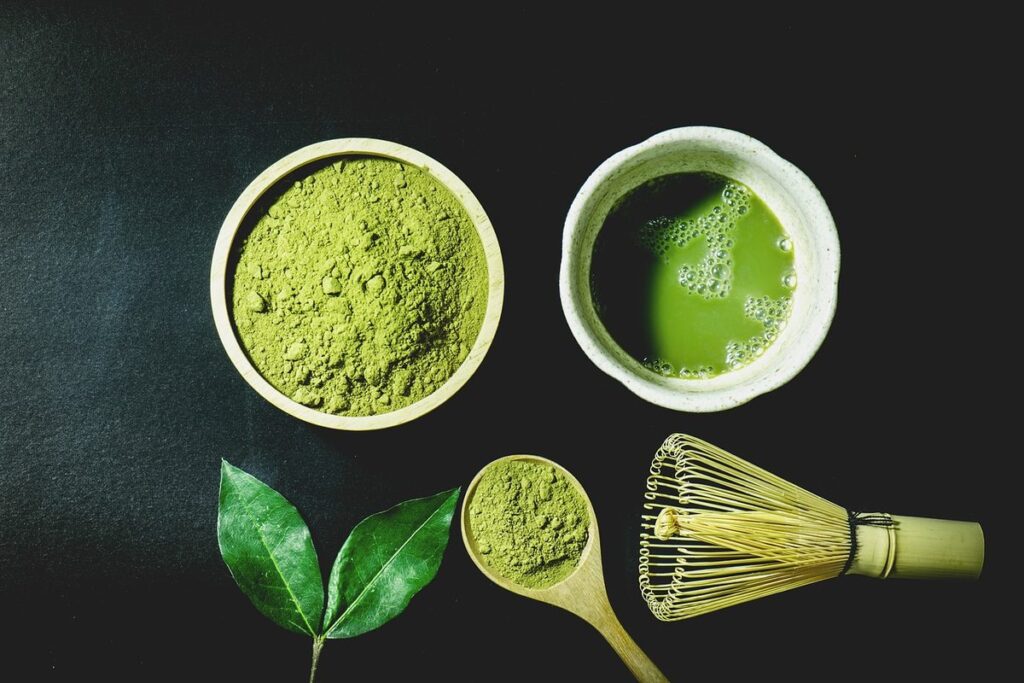 Matcha-Pulver, aufgeschlagener Matcha, Teeblätter, Bambuslöffel und Bambusbesen auf dunklem Untergrund