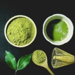 Matcha-Pulver, aufgeschlagener Matcha, Teeblätter, Bambuslöffel und Bambusbesen auf dunklem Untergrund